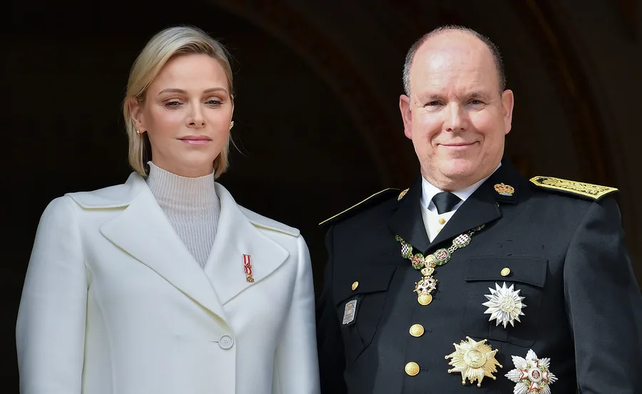A woman in a white coat stands beside a man in a decorated black uniform, both smiling slightly.