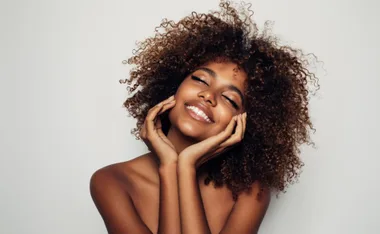 Smiling woman with curly hair enjoying skincare, resting hands on face against a plain background.