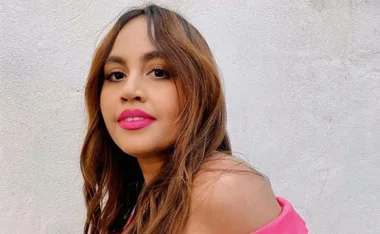 A woman with long brown hair, pink lipstick, and a pink top, smiling against a white wall background.