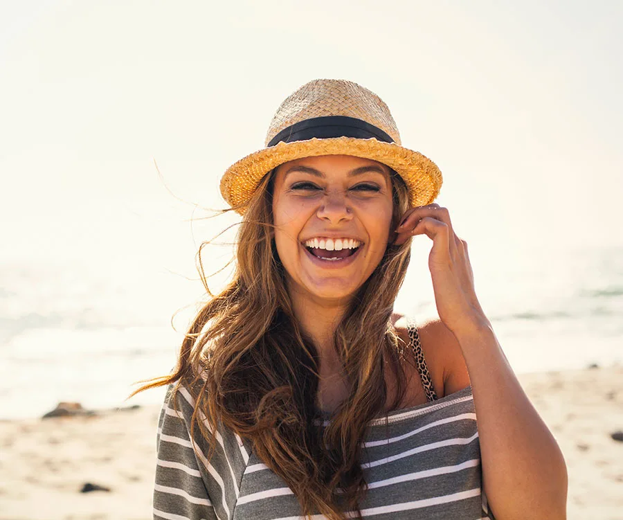 Smiling woman in a straw hat on a sunny beach, wearing a striped shirt.