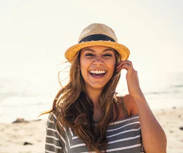 Smiling woman in a straw hat on a sunny beach, wearing a striped shirt.
