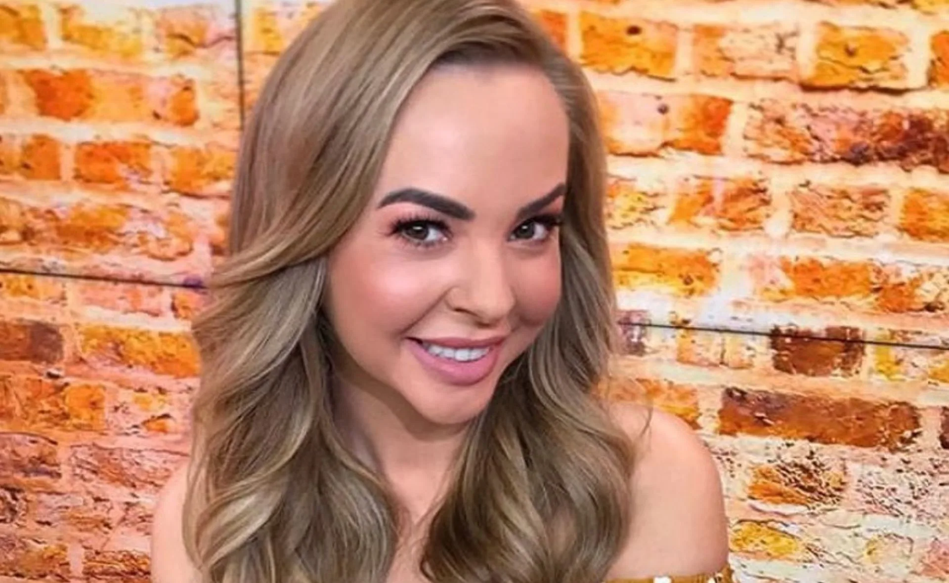Blonde woman smiling in front of a brick wall backdrop.