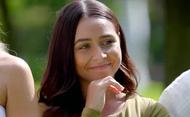 A woman with long brown hair smiles softly, outdoors with greenery in the background.