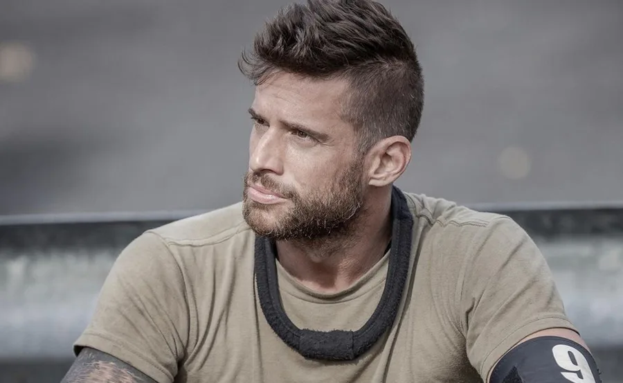 A man with short hair and beard wearing a tan shirt, sitting outdoors, looking pensively to the side.