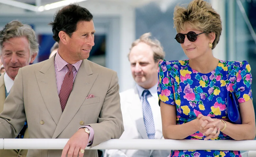 Princess Diana in a vibrant floral dress and sunglasses, standing next to a man in a beige suit, engaged in conversation.