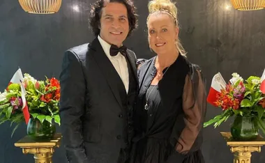 A couple dressed elegantly stands together, with floral arrangements in the background.