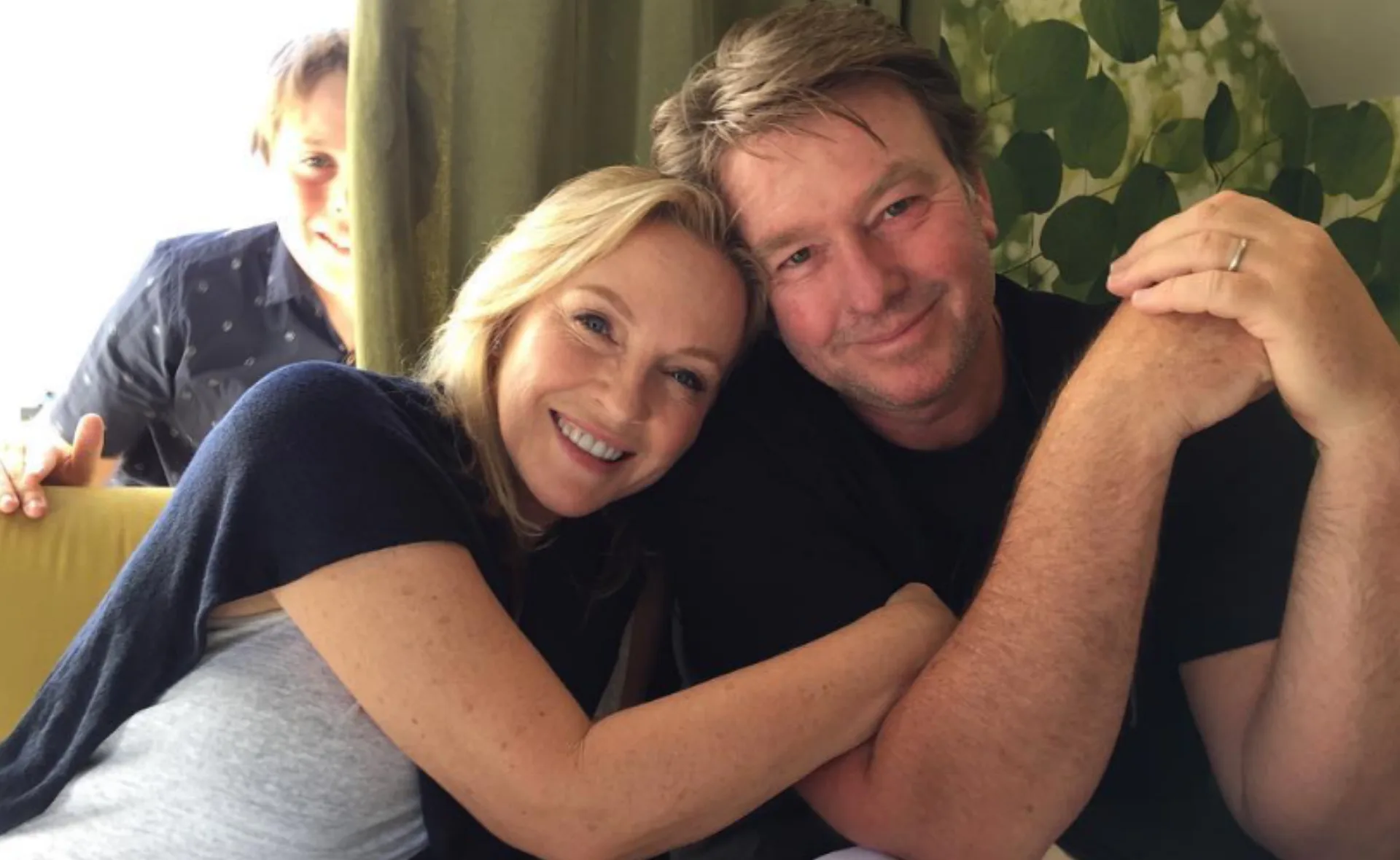 Couple sitting closely, smiling at the camera, with a person playfully photobombing from behind.