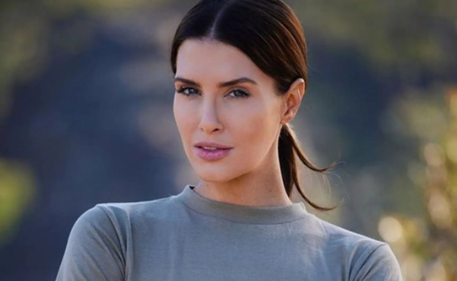 Woman with dark hair tied back, wearing a gray top, looking confidently at the camera, blurred nature background.