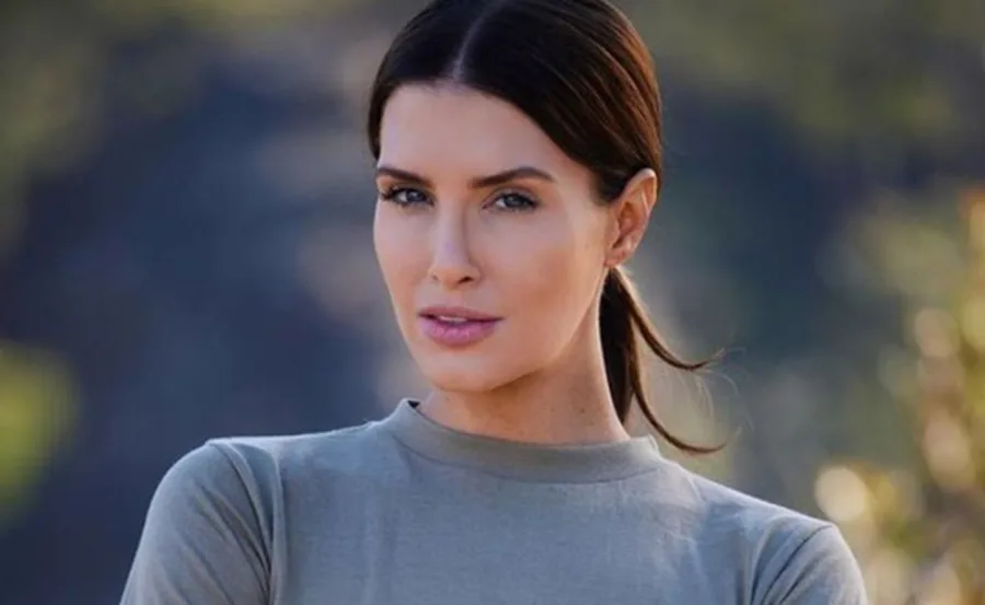 Woman with dark hair tied back, wearing a gray top, looking confidently at the camera, blurred nature background.