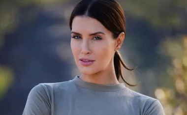 Woman with dark hair tied back, wearing a gray top, looking confidently at the camera, blurred nature background.