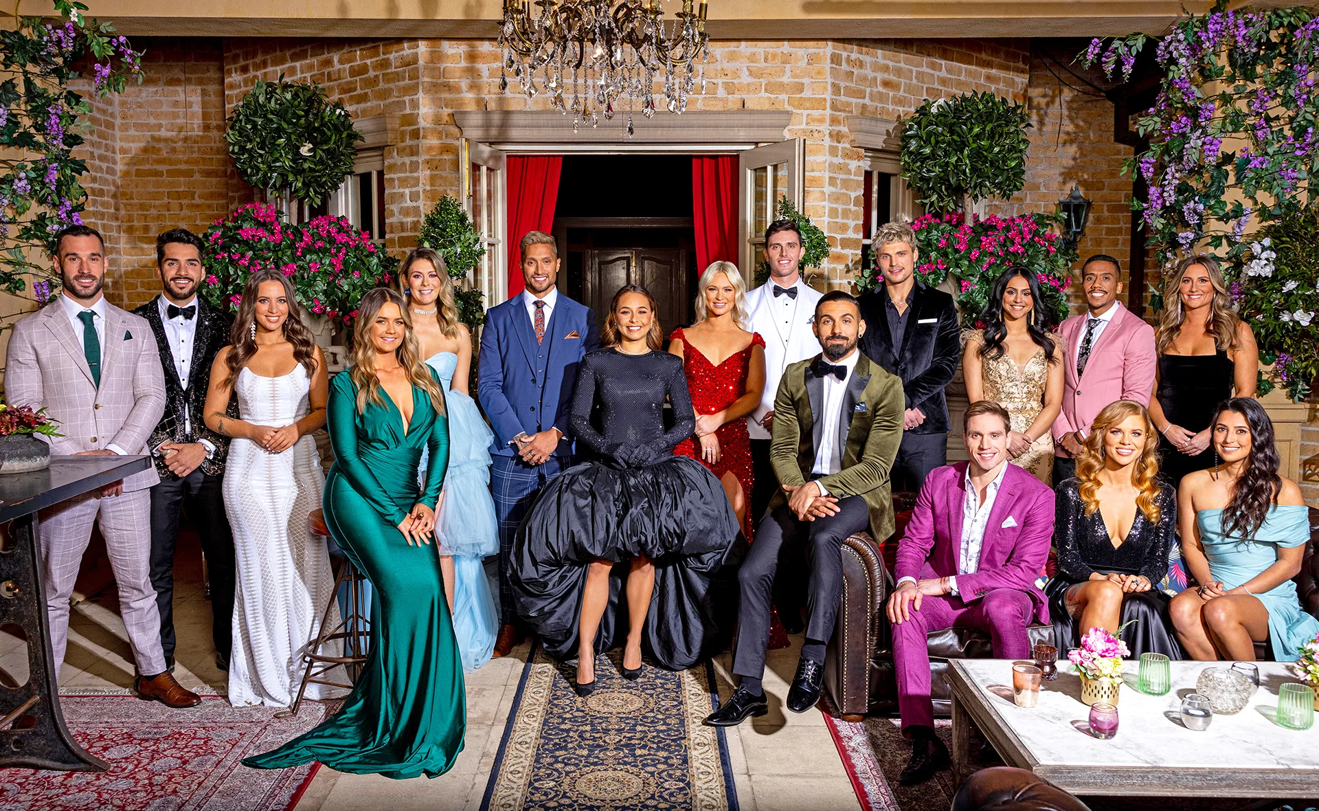 Group photo of The Bachelorette 2021 cast in formal attire, standing and sitting in an ornate room with floral decorations.