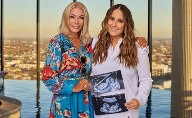 Two women smiling and holding ultrasound images, standing in front of a large window with a cityscape in the background.