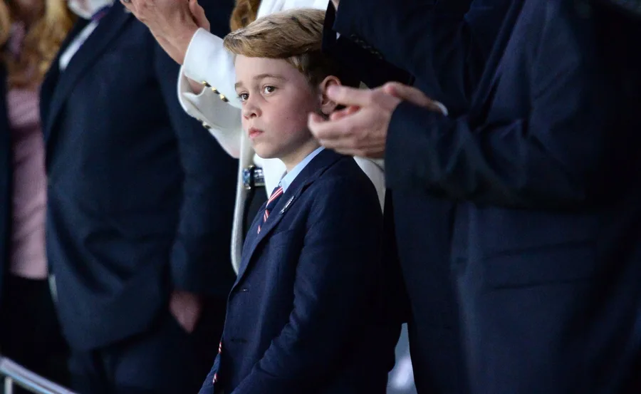 A young boy in a suit and tie stands among adults, looking thoughtful, at an event with people clapping around him.