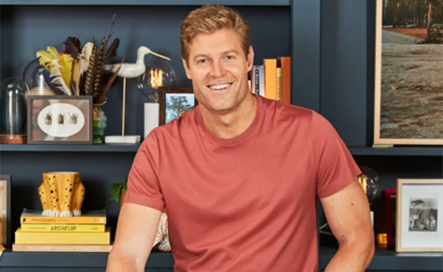 A person in a red shirt smiling in front of a blue bookshelf with books and decorative items.