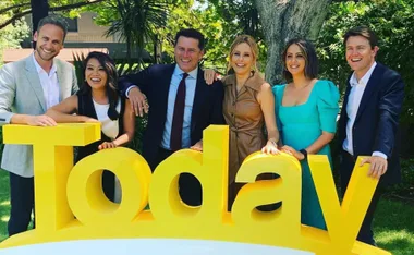 Group of six people smiling outdoors, leaning on a large yellow "Today" sign.