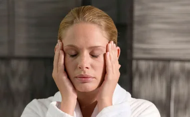Woman in a white robe gently massages her temples with her eyes closed.