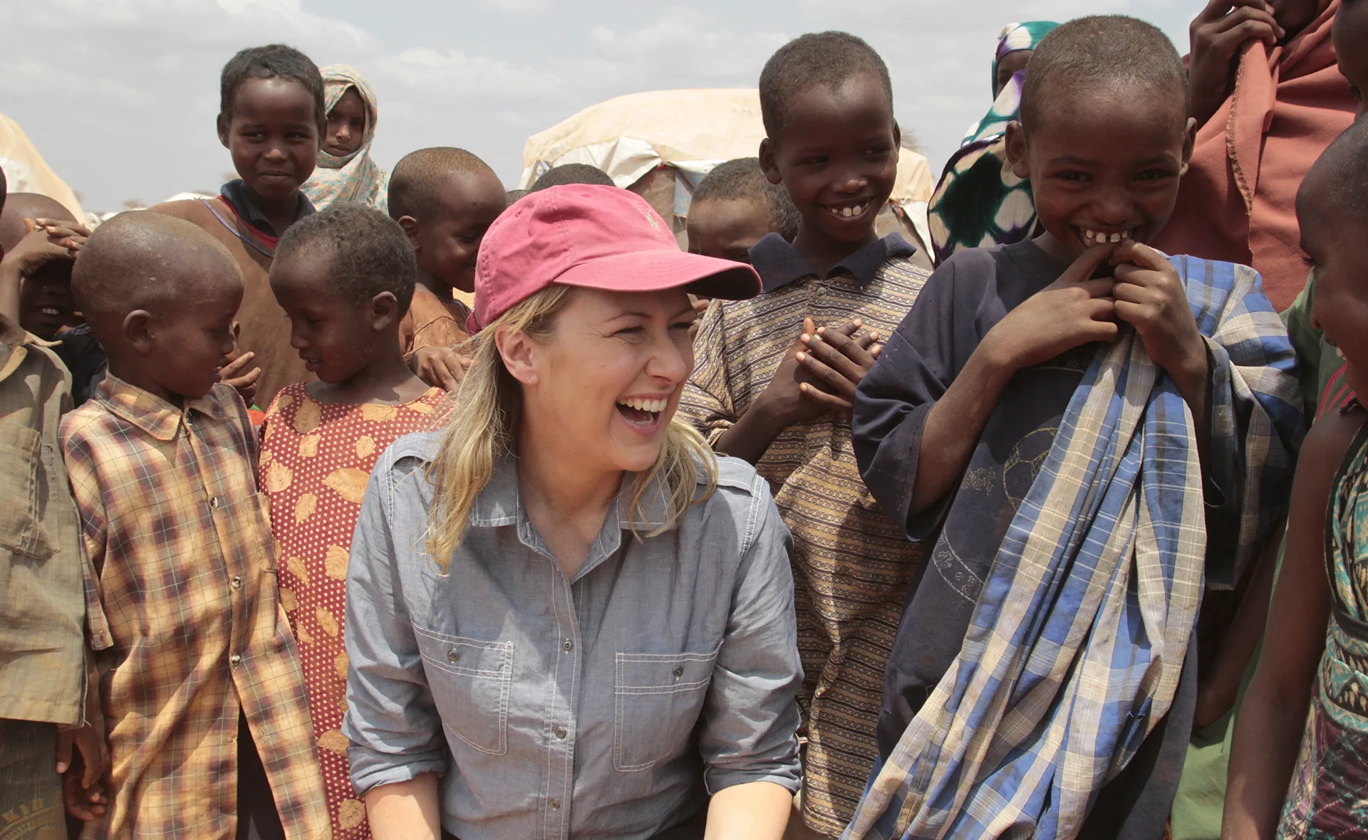 A smiling woman in a cap surrounded by happy children outdoors, engaging in friendly interaction.