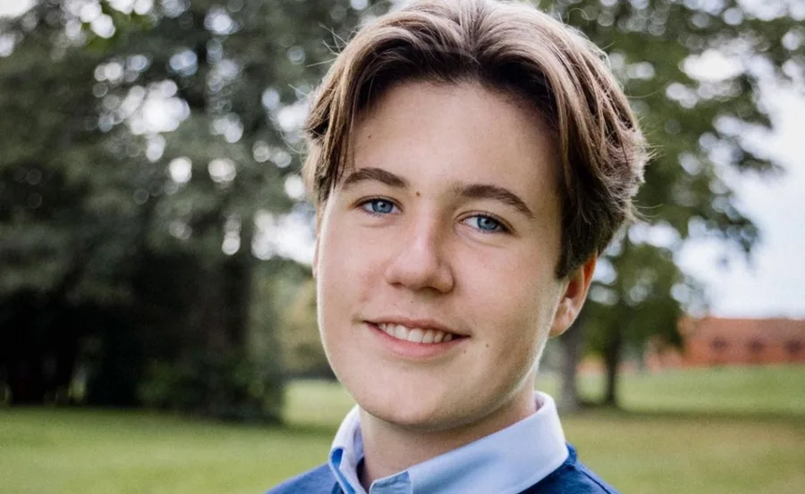 Prince Christian smiling outdoors, wearing a blue sweater and white shirt, trees blurred in the background.