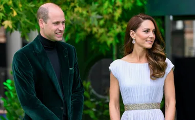 A man in a green velvet suit and a woman in a white dress stand together outdoors, surrounded by greenery.