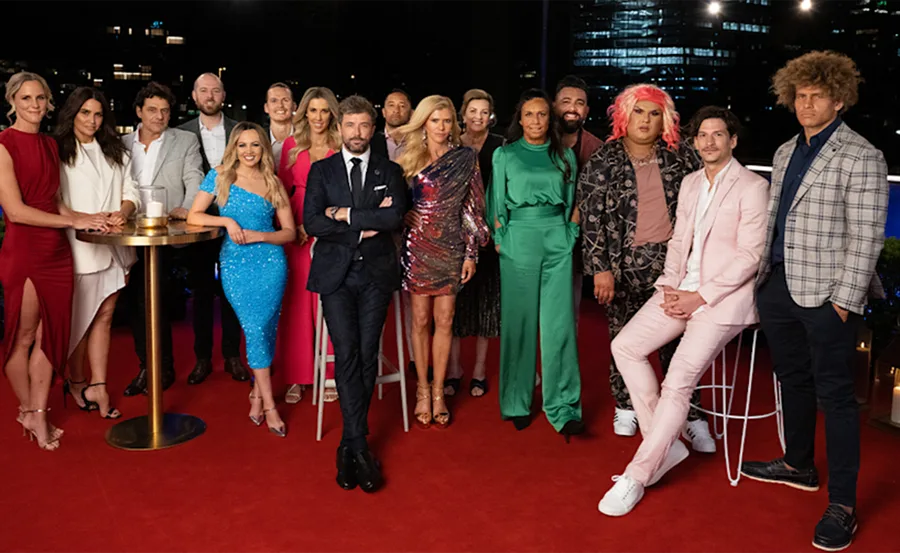 Group of people dressed in formal attire standing on a red carpet at night, with city lights in the background.