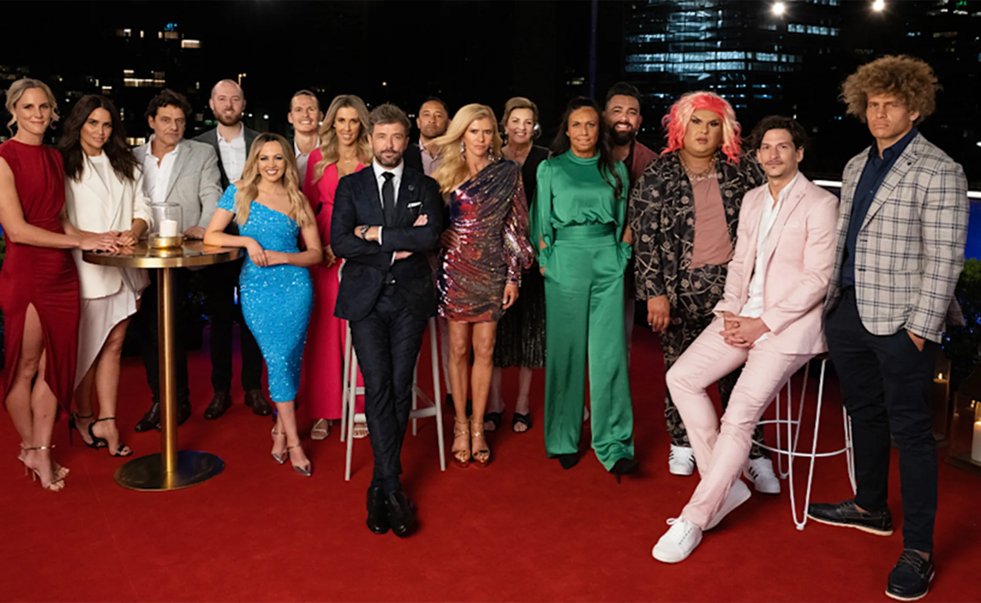 Group of people dressed in formal attire standing on a red carpet at night, with city lights in the background.