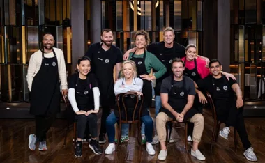 "Eight contestants smiling in black aprons on a wooden floor, part of a cooking show setting."