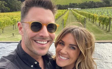 A smiling couple taking a selfie in front of lush vineyard rows on a sunny day.