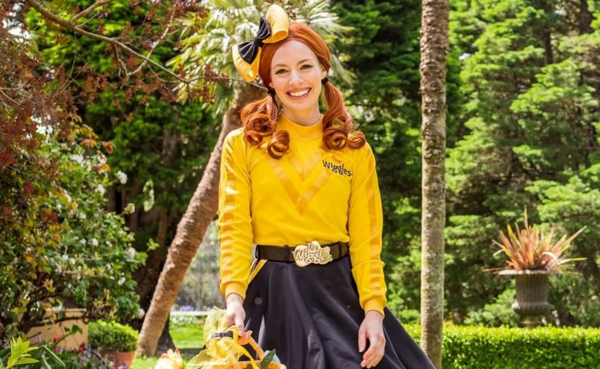 Emma Watkins from The Wiggles, wearing a yellow and black outfit, smiling in a garden setting.