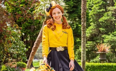 Emma Watkins from The Wiggles, wearing a yellow and black outfit, smiling in a garden setting.
