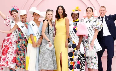 Group of eight people in vibrant outfits, smiling in front of a pink backdrop at the Melbourne Cup fashion event.