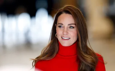 A person with long brown hair wearing a red turtleneck, smiling indoors.