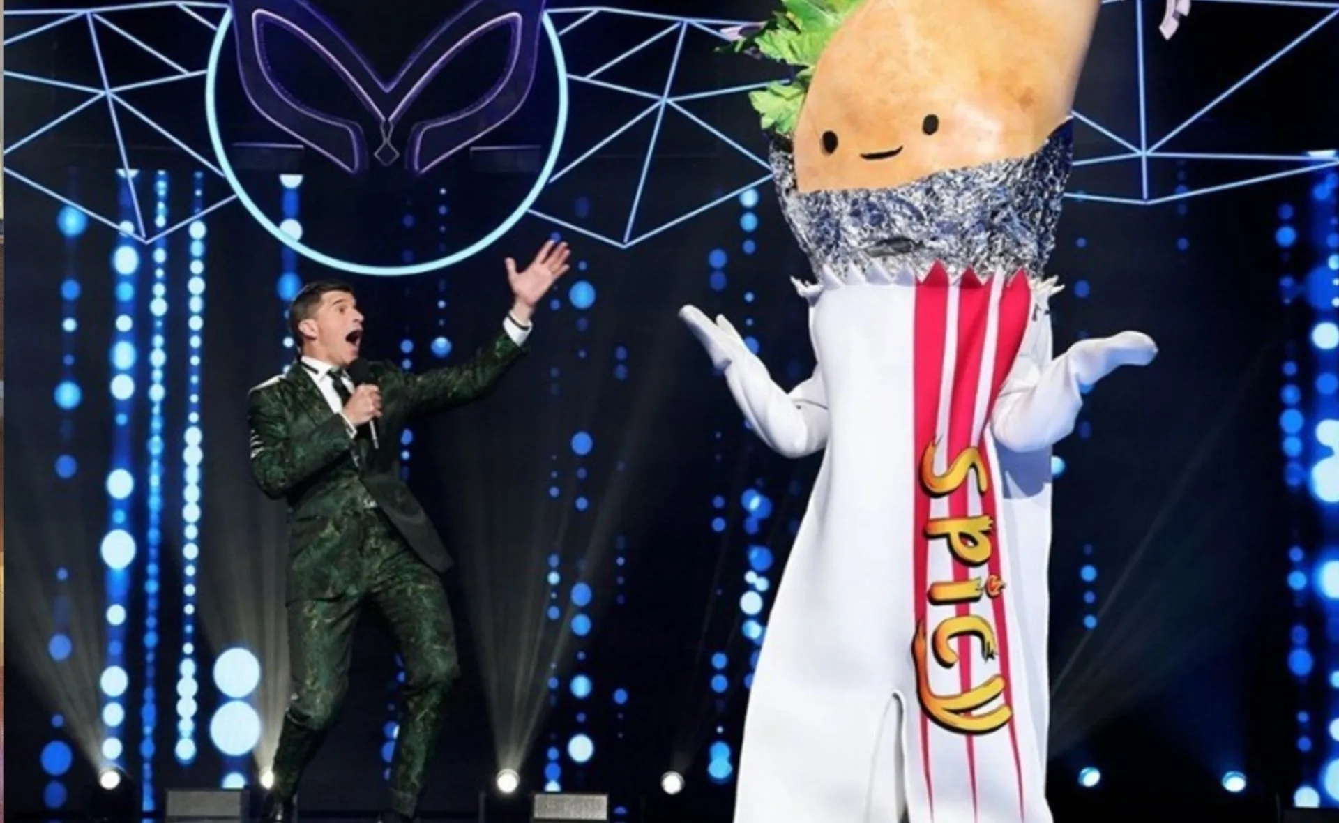 Host in a patterned suit with a smiling taco costume character labeled "Spicy" on a brightly lit stage.