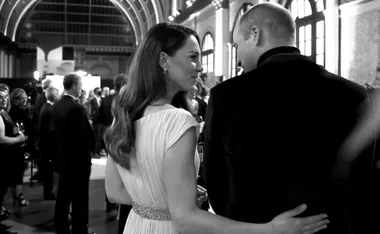 Couple in formal attire embraces, chatting at a social event in a grand hall.