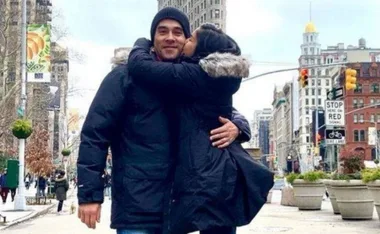A woman kisses a smiling man on the cheek in a city street with historical architecture in the background.