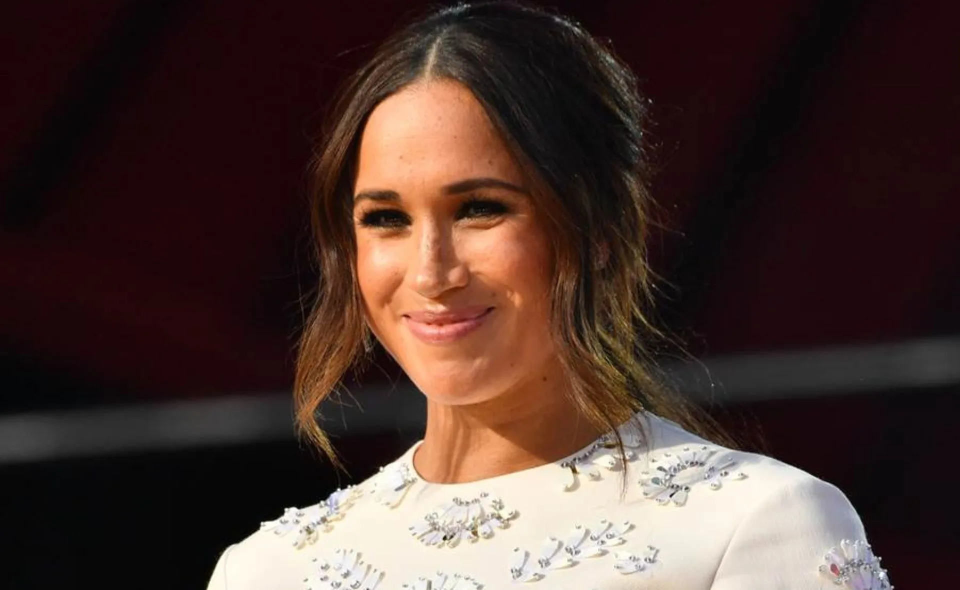 Meghan Markle smiling, wearing a white embellished top, with her hair styled in a loose updo.