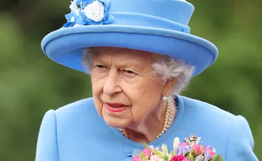 Elderly woman in a blue outfit and hat, holding a bouquet of flowers, smiling.