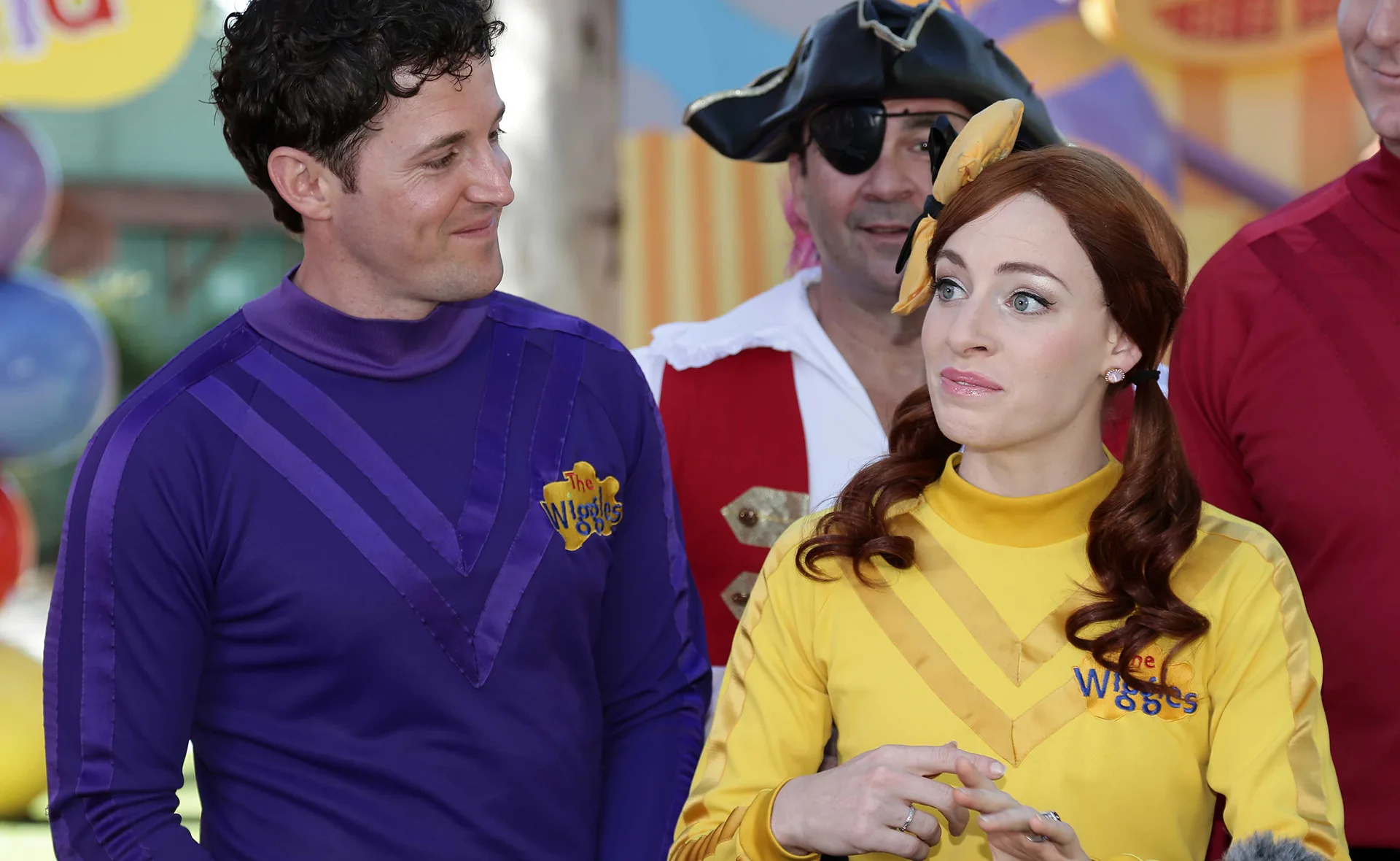 Members of The Wiggles in costume, including one in a purple suit and one in a yellow suit, interacting with each other.