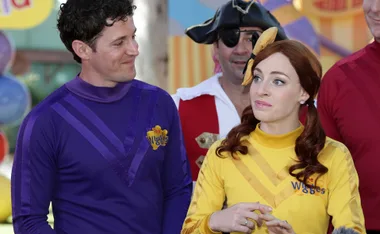 Members of The Wiggles in costume, including one in a purple suit and one in a yellow suit, interacting with each other.