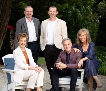 Group photo of five people from the show "Neighbours" sitting and standing outdoors, smiling at the camera.