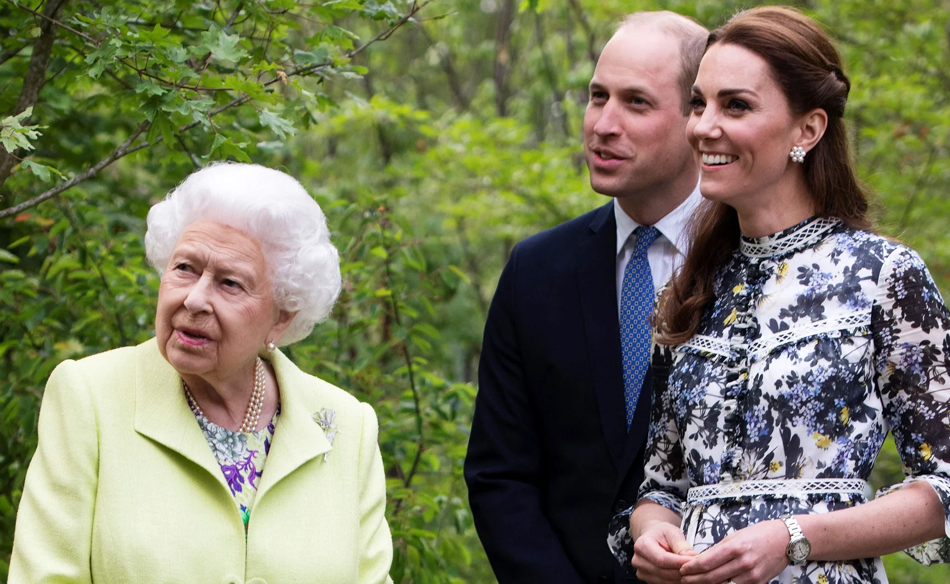 Three people standing outdoors, two smiling, with greenery in the background.