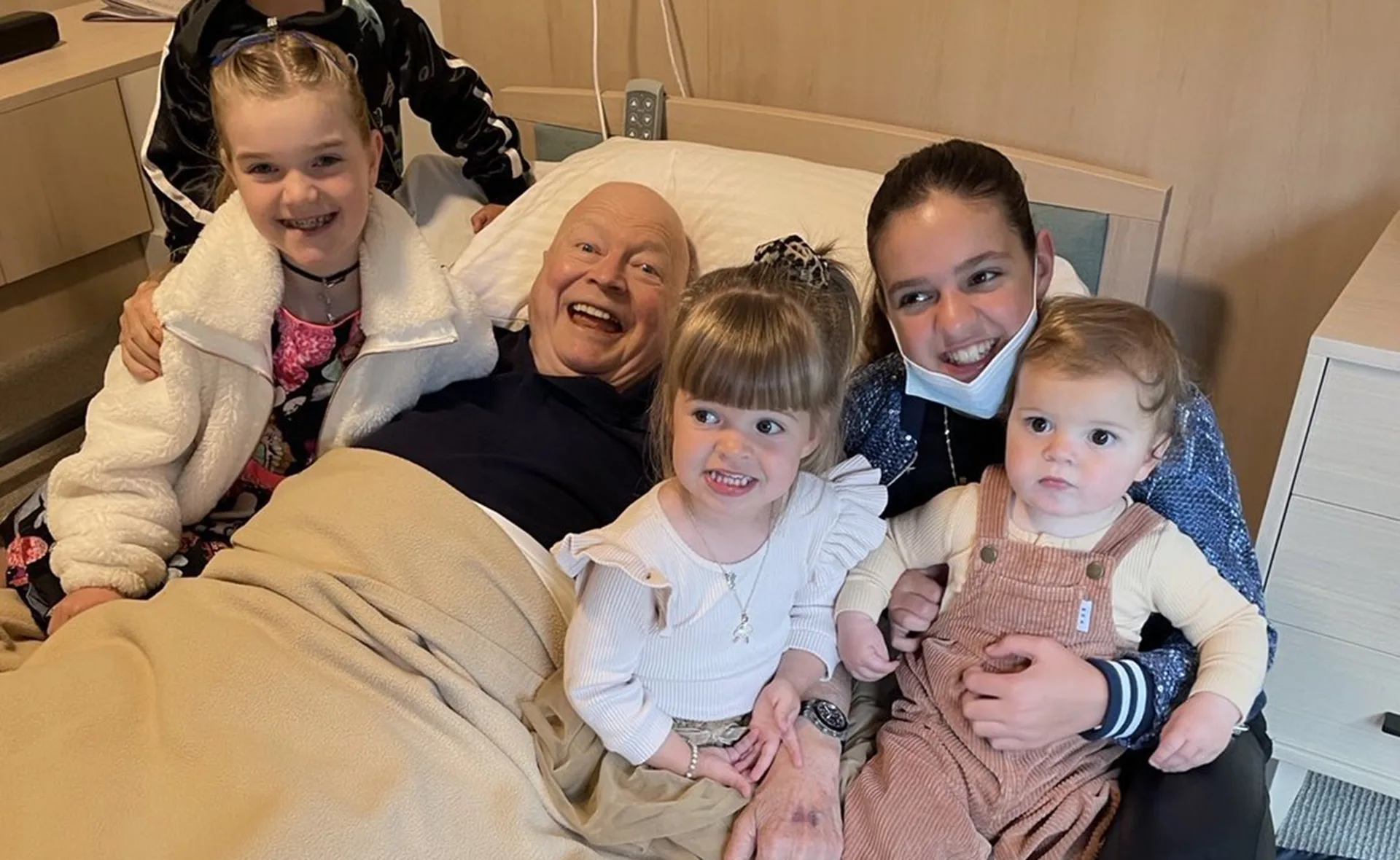 An elderly man is in a hospital bed surrounded by smiling children and a young adult, creating a joyful atmosphere.