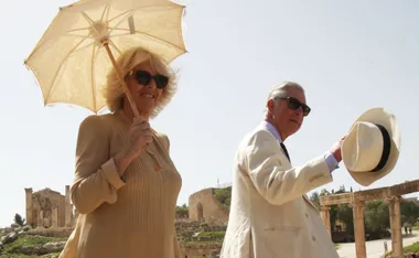Couple in light attire, one holding a parasol, the other tipping a hat, explore ancient ruins under a clear sky.