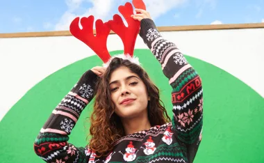 Young woman in festive sweater with red antler headband, standing outdoors against a green and white background.