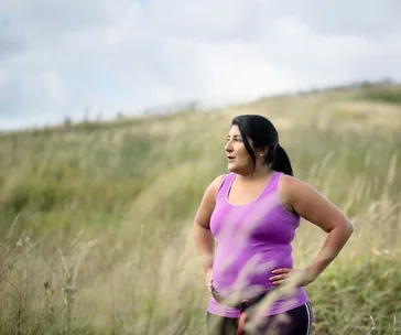 Woman in a purple tank top standing in a grassy field, looking to the side.