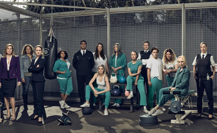 A group of characters from the TV show "Wentworth" stands in a prison yard, some in uniforms, others in teal inmate clothing.