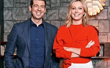 A man in a blue suit and a woman in a red blouse smiling, standing in front of a textured wall indoors.