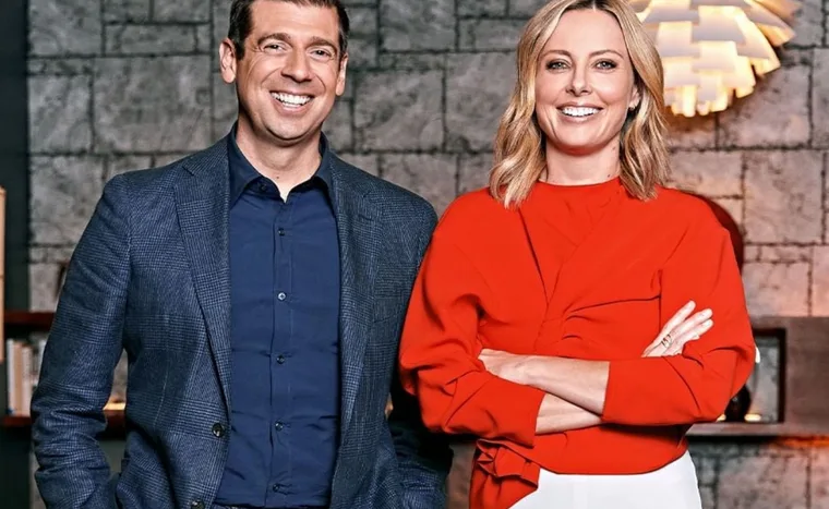 A man in a blue suit and a woman in a red blouse smiling, standing in front of a textured wall indoors.