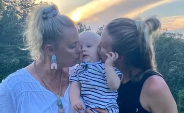 Two women kissing a baby on both cheeks at sunset, surrounded by trees.