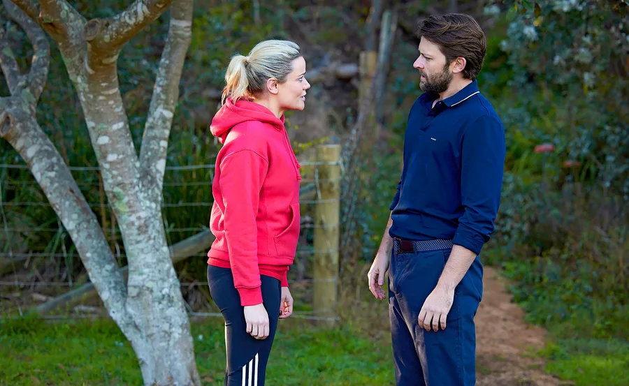 Two people stand outdoors, facing each other, with trees and a path in the background.