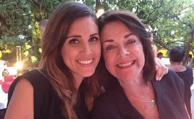 Two smiling women, close together, outdoors at an event with greenery in the background.